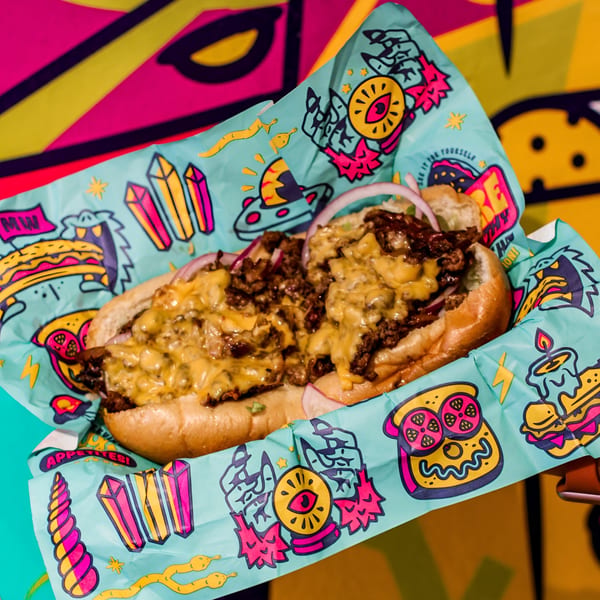 Cheesesteak sandwich with melted cheese and red onions, served in colorful, illustrated paper.