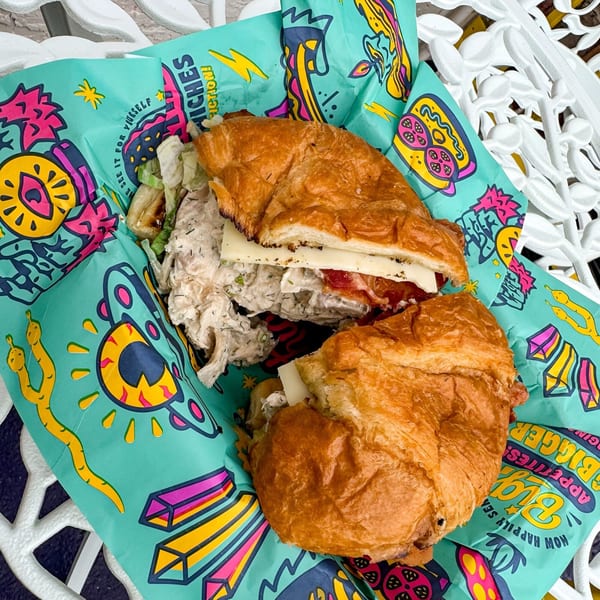 A chicken salad sandwich with cheese and bacon on a croissant, wrapped in colorful paper.