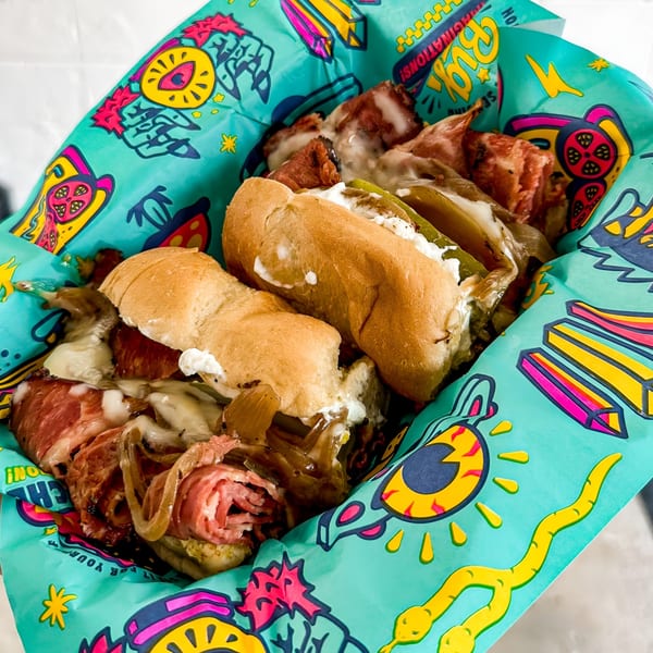 Two hearty sandwiches with meat, cheese, and vegetables in a vibrant, patterned paper basket.