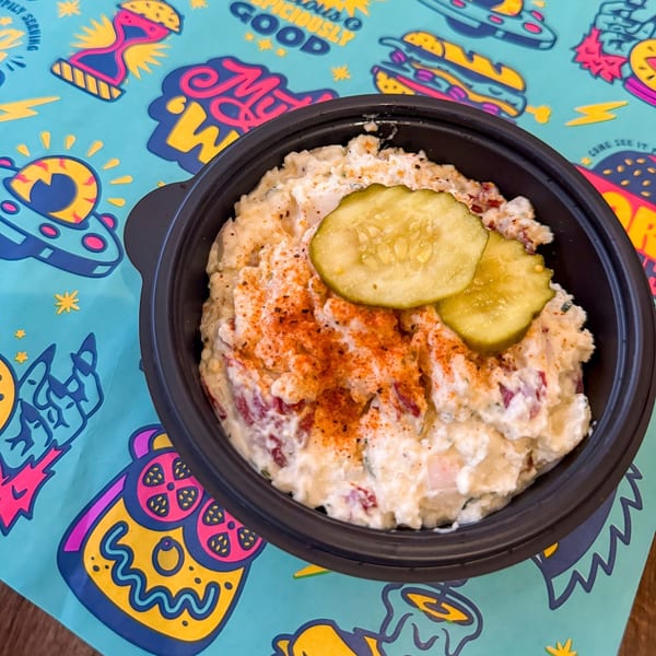 Creamy potato salad with paprika and two pickle slices in a black bowl on a colorful placemat.