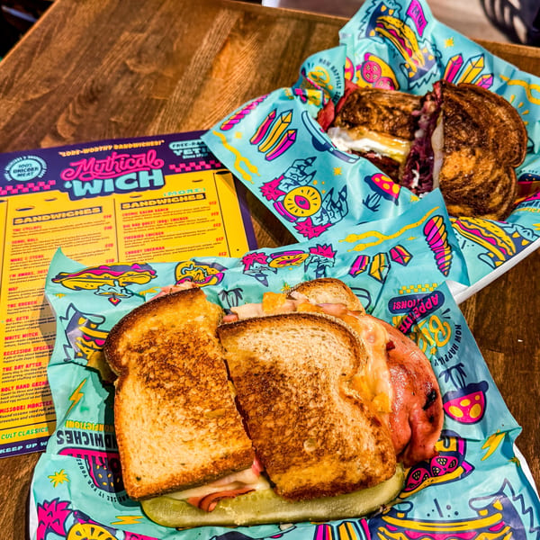Two sandwiches, one cut in half, on colorful Mythical 'Wich branded paper, with a menu beside them.
