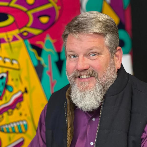 Smiling man with a gray beard and purple shirt in front of a colorful, abstract background.