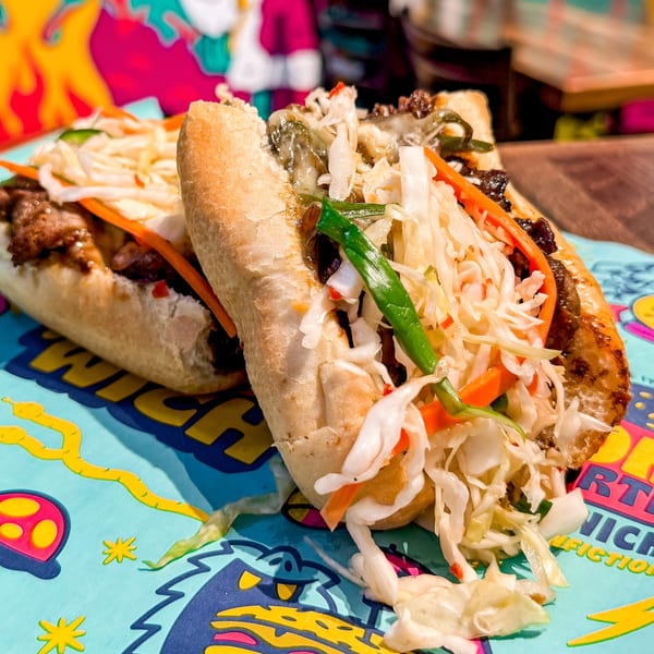 Two banh mi sandwiches with meat, pickled vegetables, and herbs on a colorful placemat.