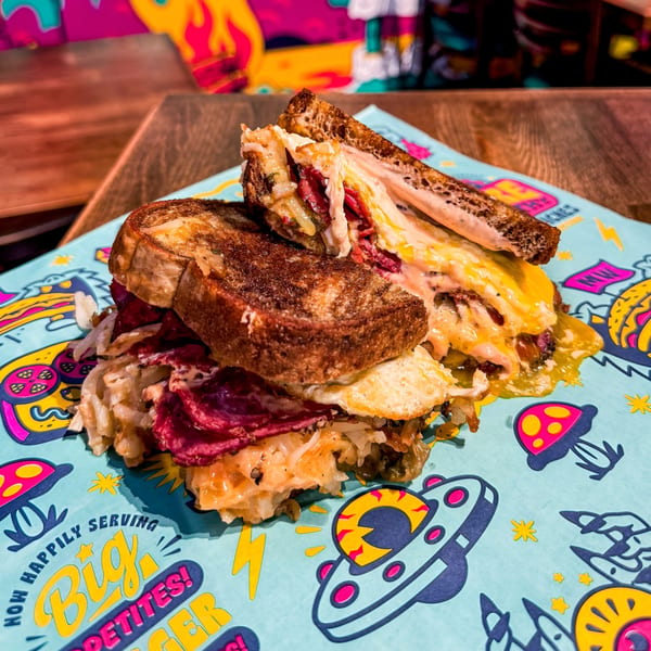 A grilled sandwich with pastrami, melted cheese, and sauerkraut on colorful, cartoon-patterned paper.
