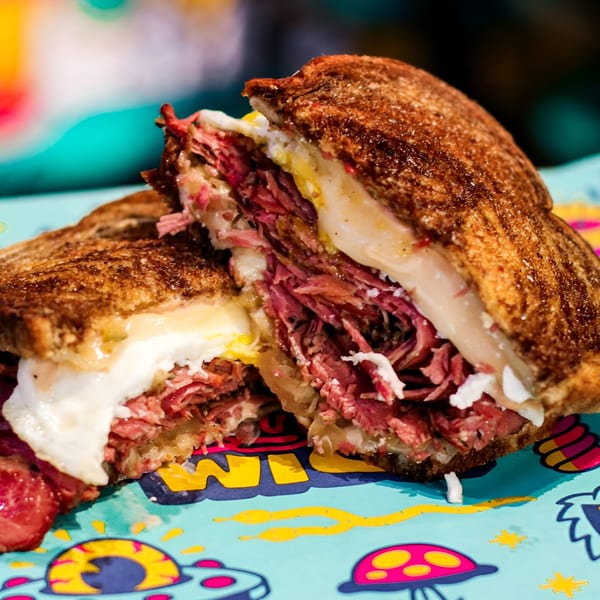A grilled sandwich with pastrami, melted cheese, and a fried egg, cut in half on a colorful placemat.
