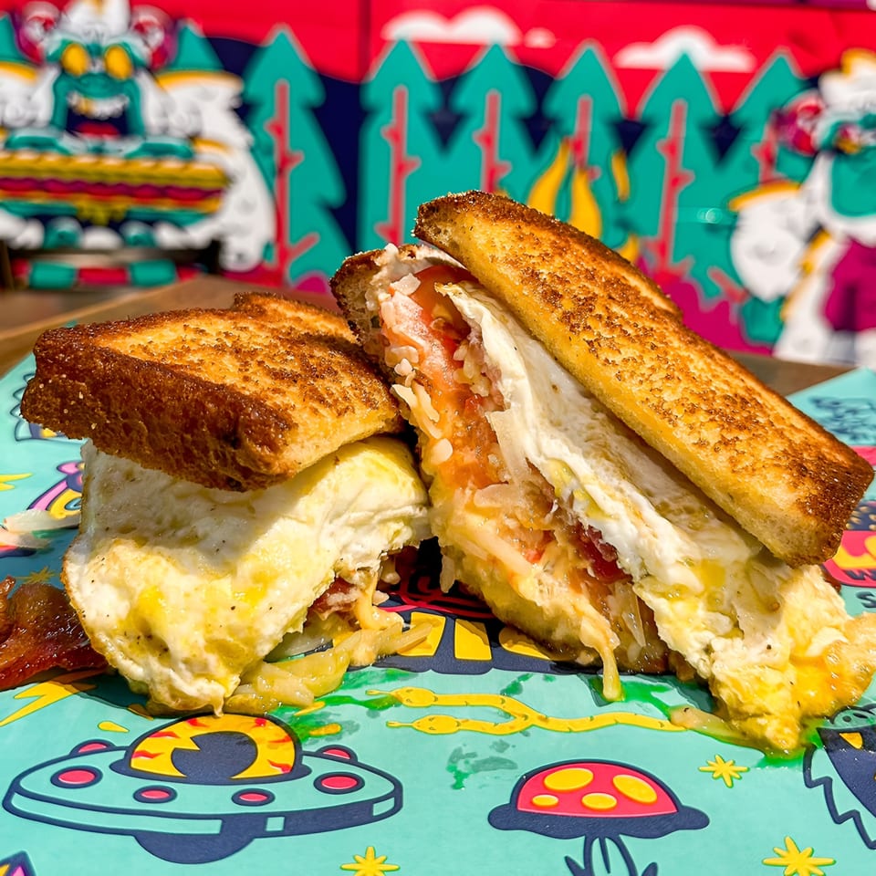 A grilled cheese sandwich with egg and bacon, cut in half, on a colorful placemat.